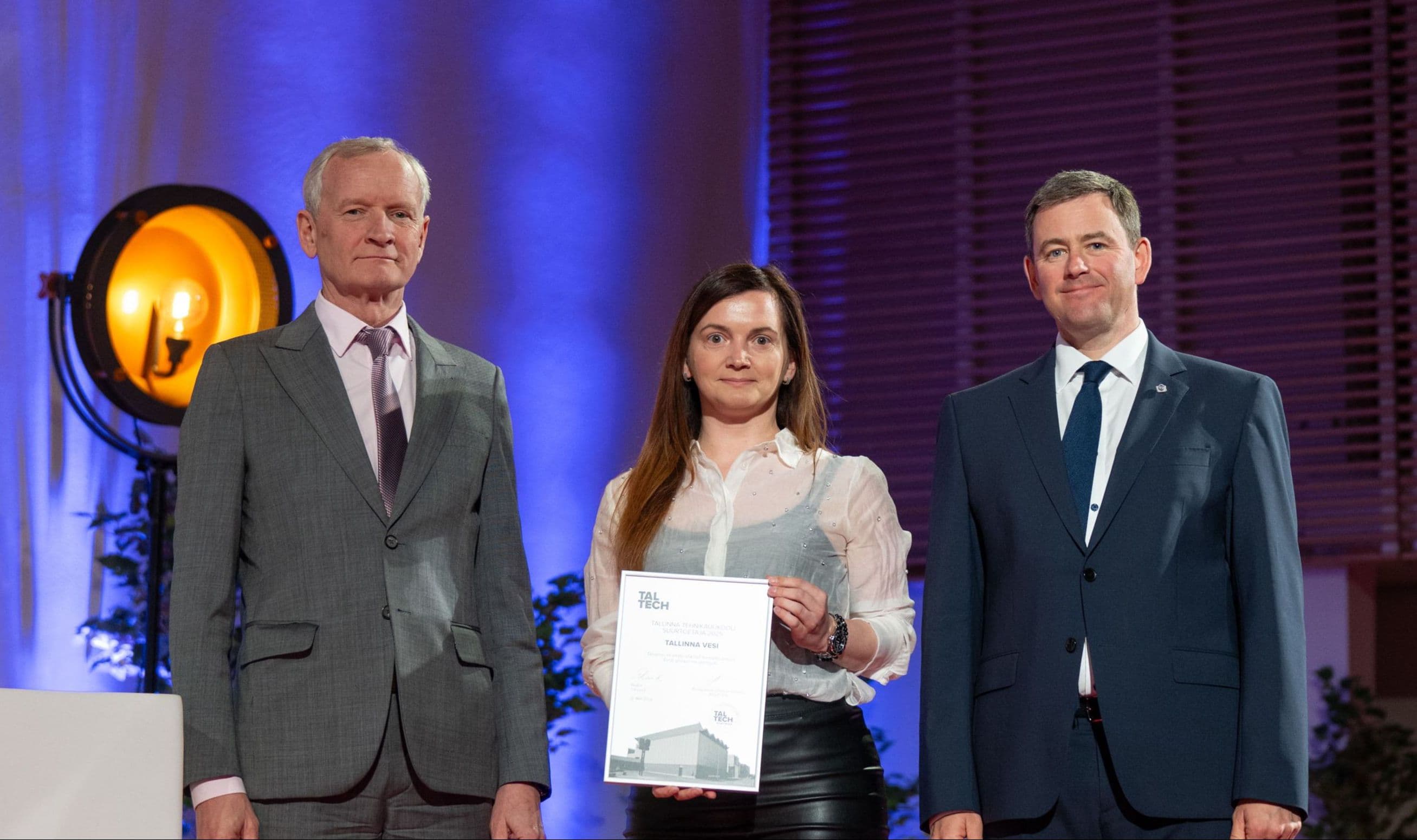 A woman receives a certificate from two men on a formal stage during an awards ceremony.