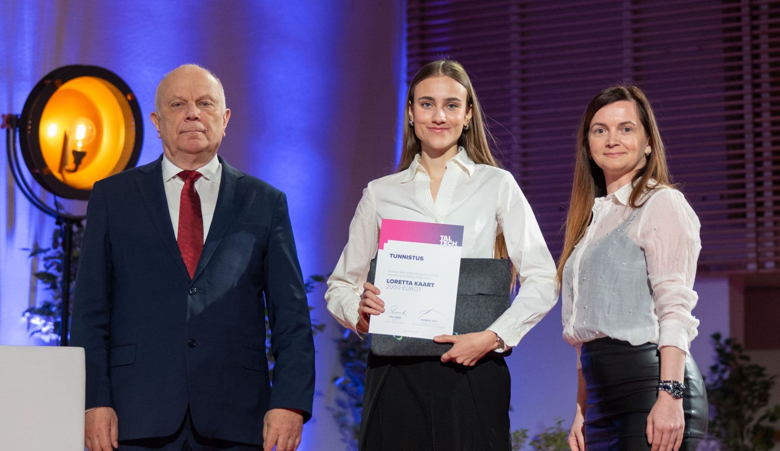 Three people on stage, a man and two women, one holding certificates.
