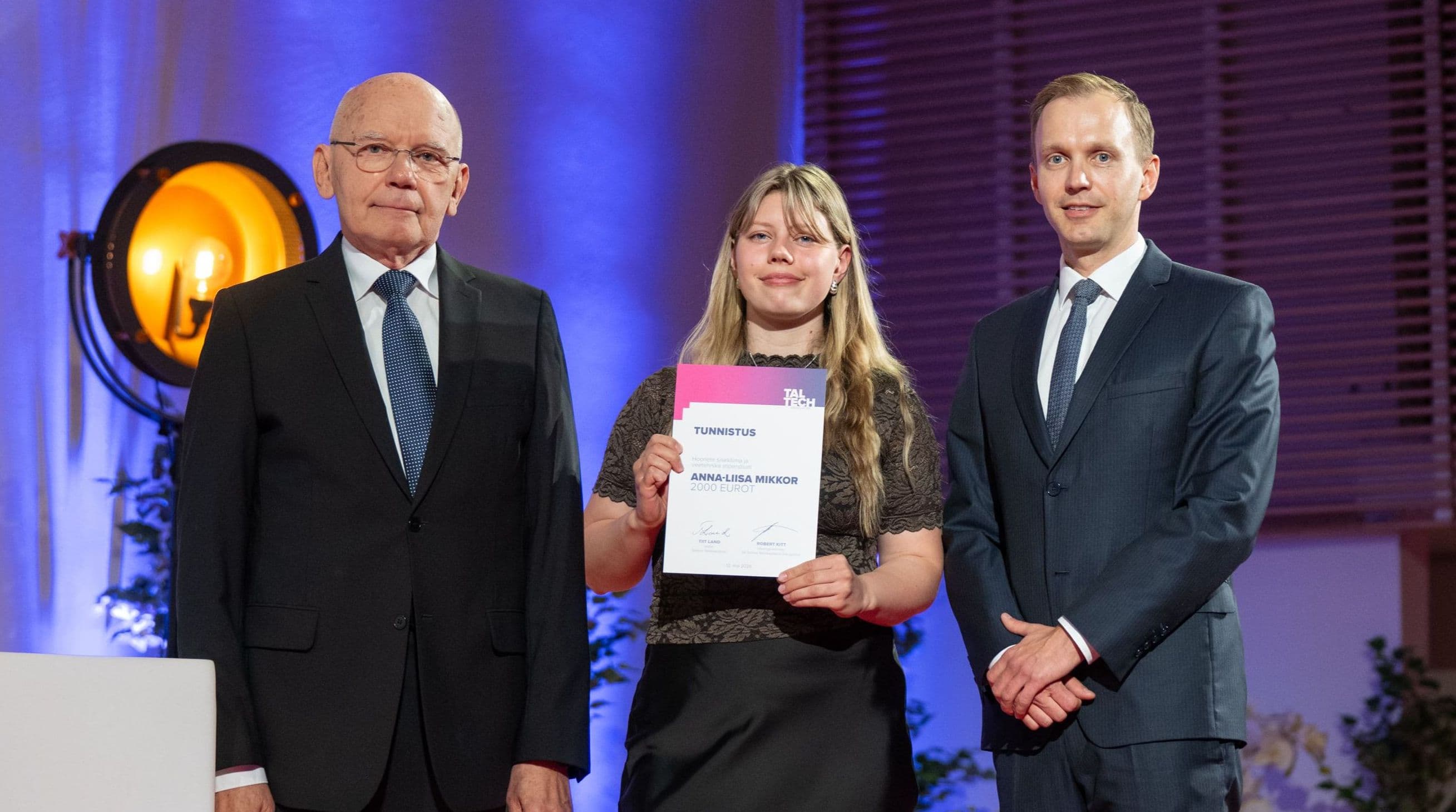 Woman receiving an award from two men on a stage at a formal ceremony.