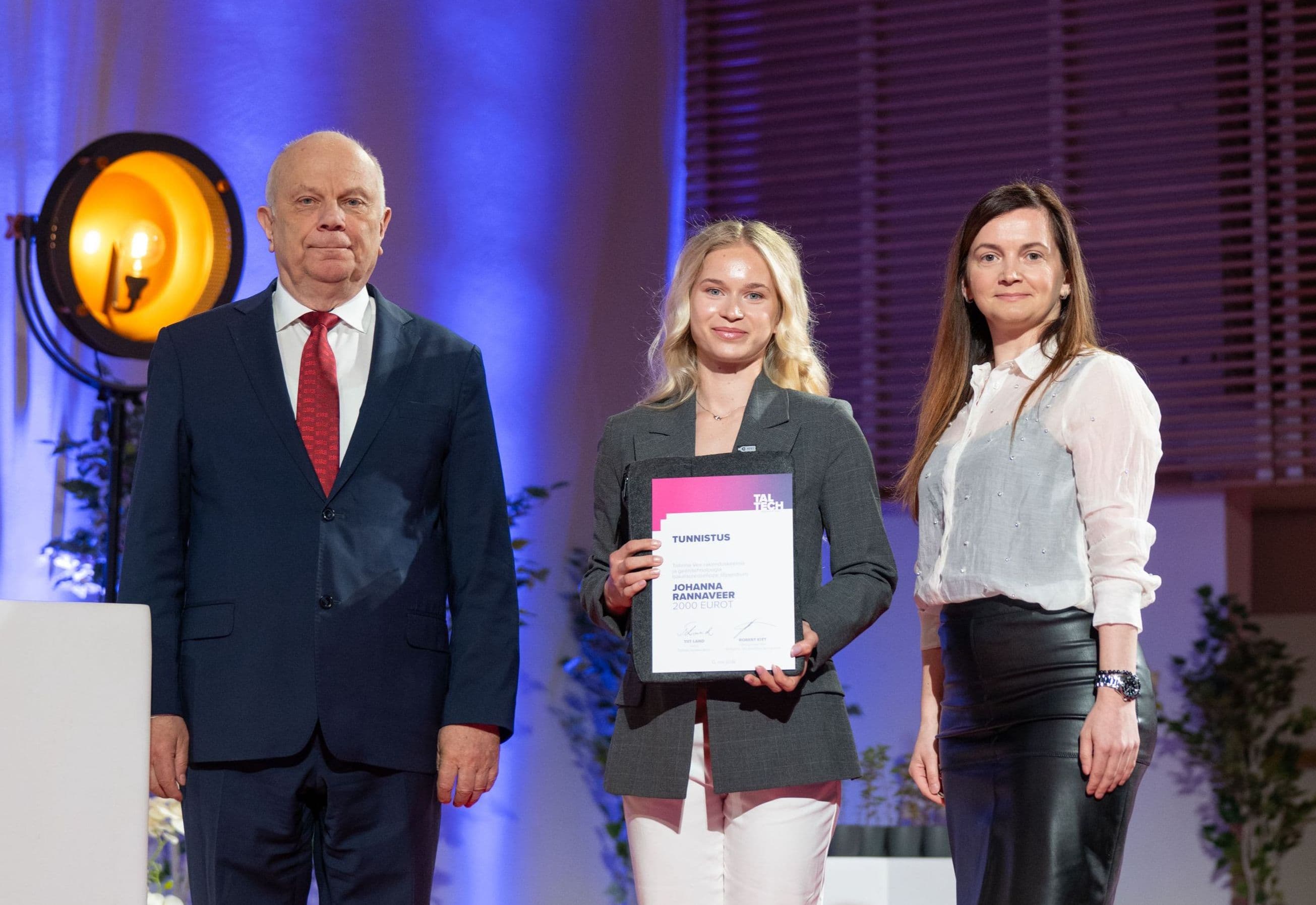 Three people on stage, a young woman in the middle holding an award certificate.