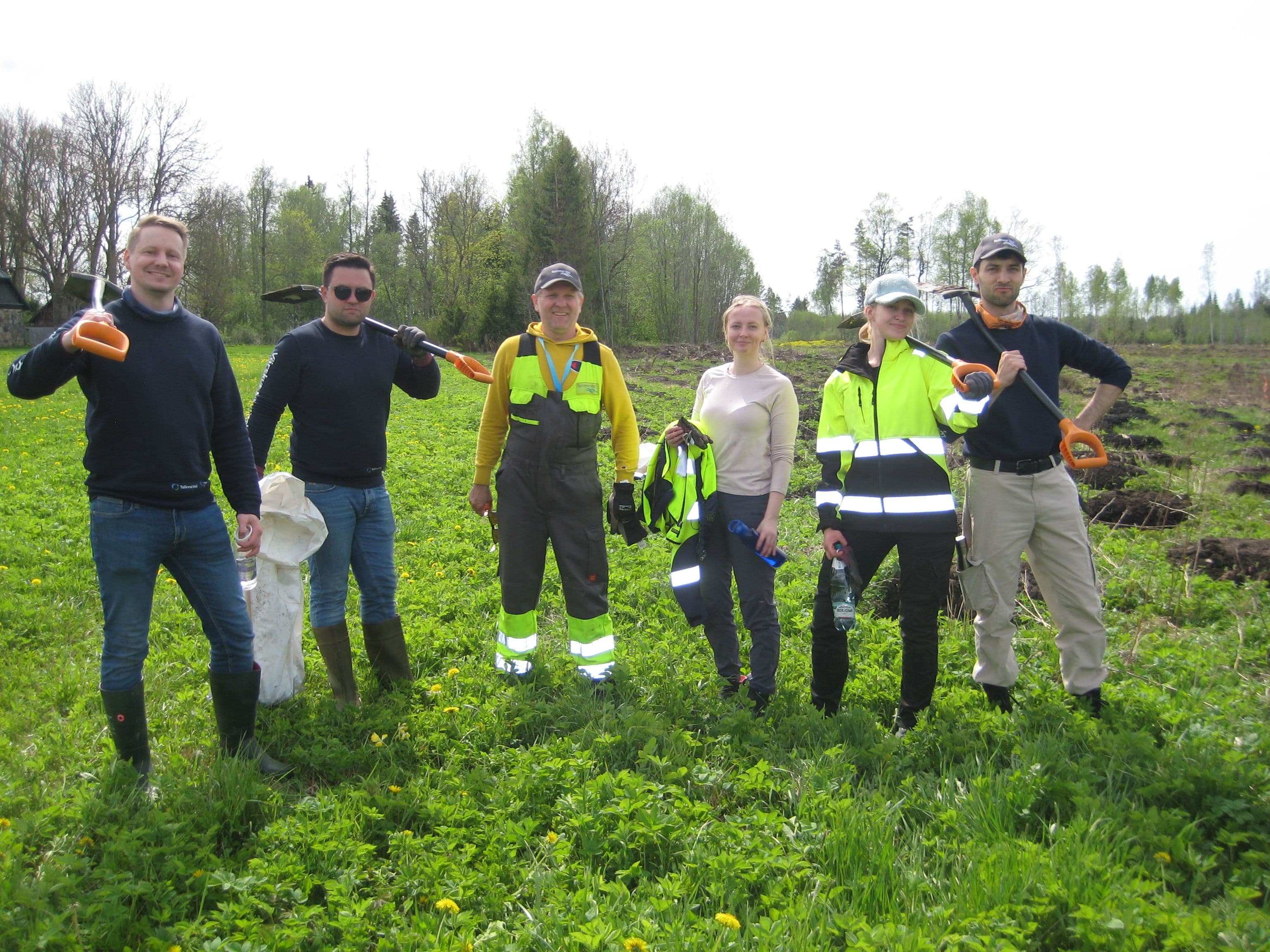 Six volunteers stand smiling in a green field, holding tools for a cleanup.