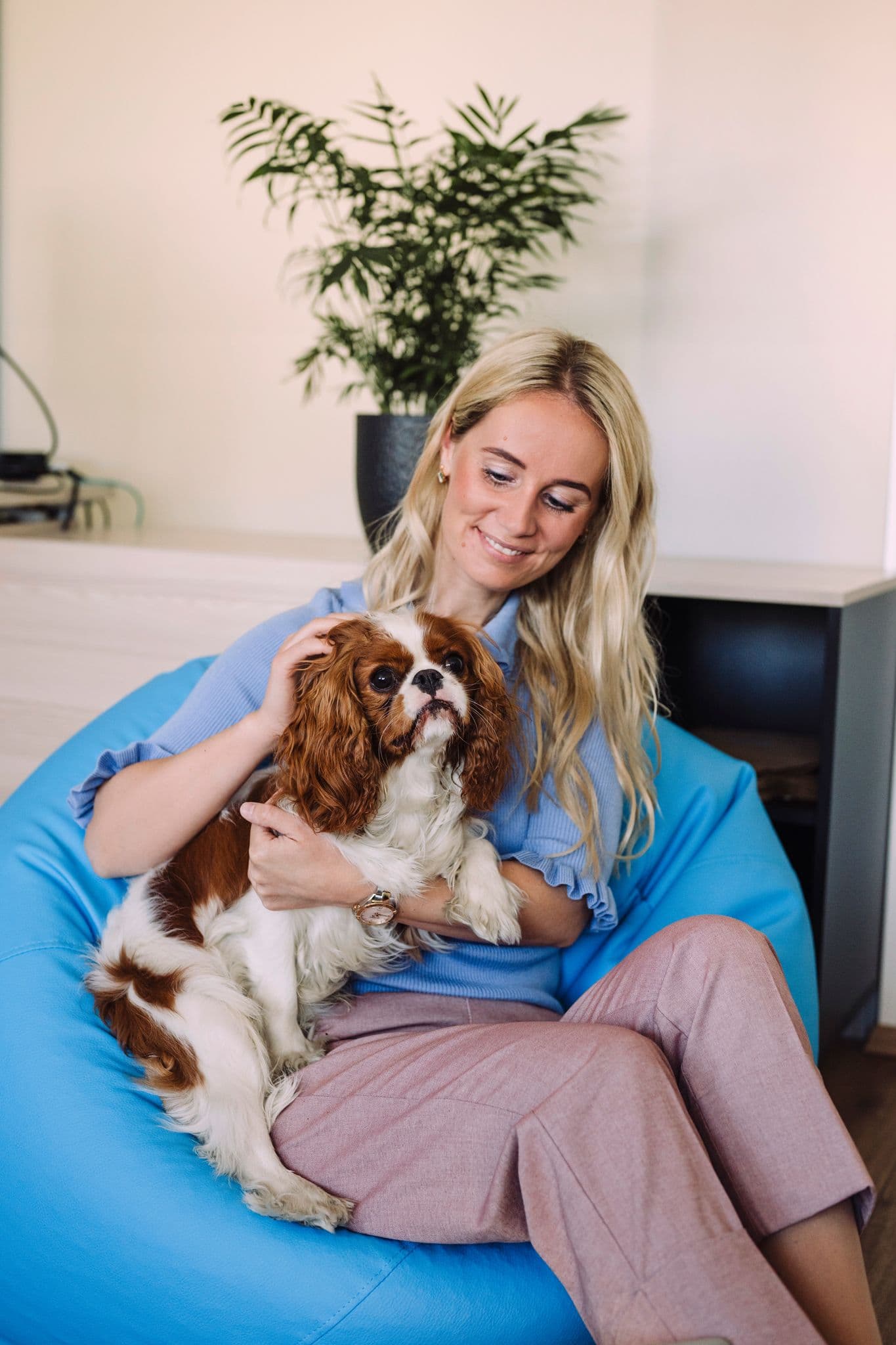 Blonde woman sitting on a blue bean bag chair, happily petting her small dog.