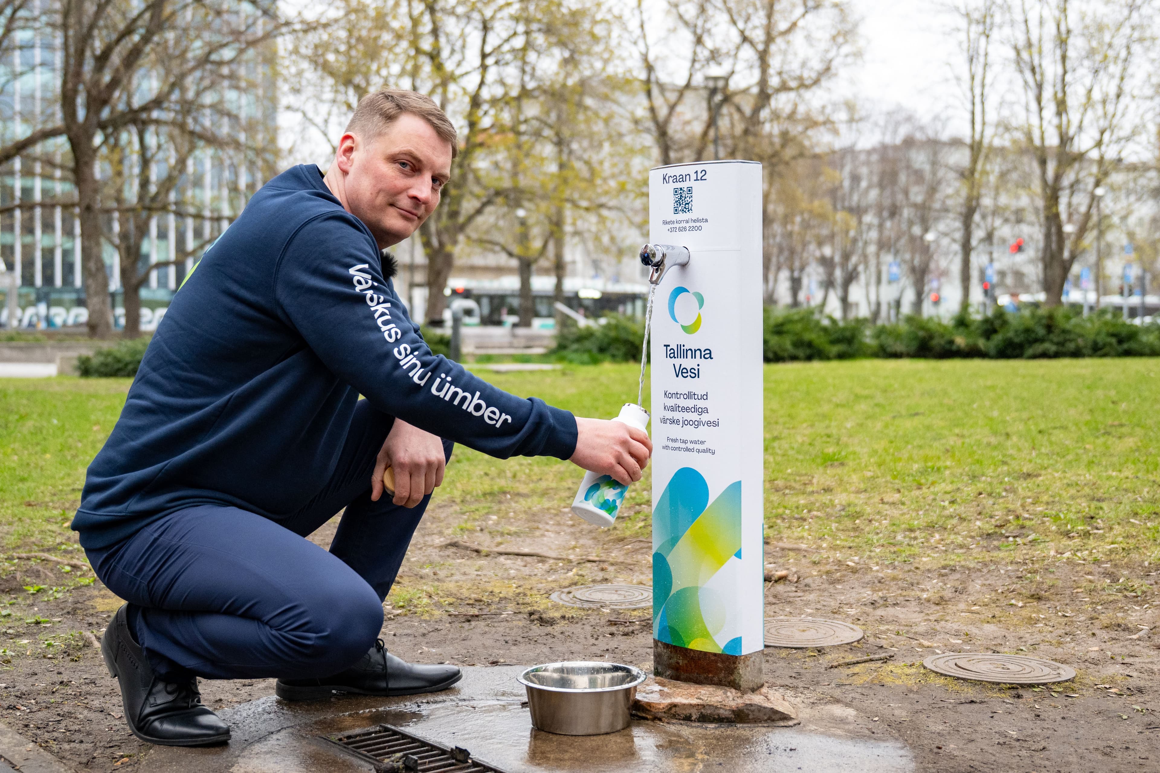 Man fills water bottle from a public dispenser with a dog bowl on the ground.