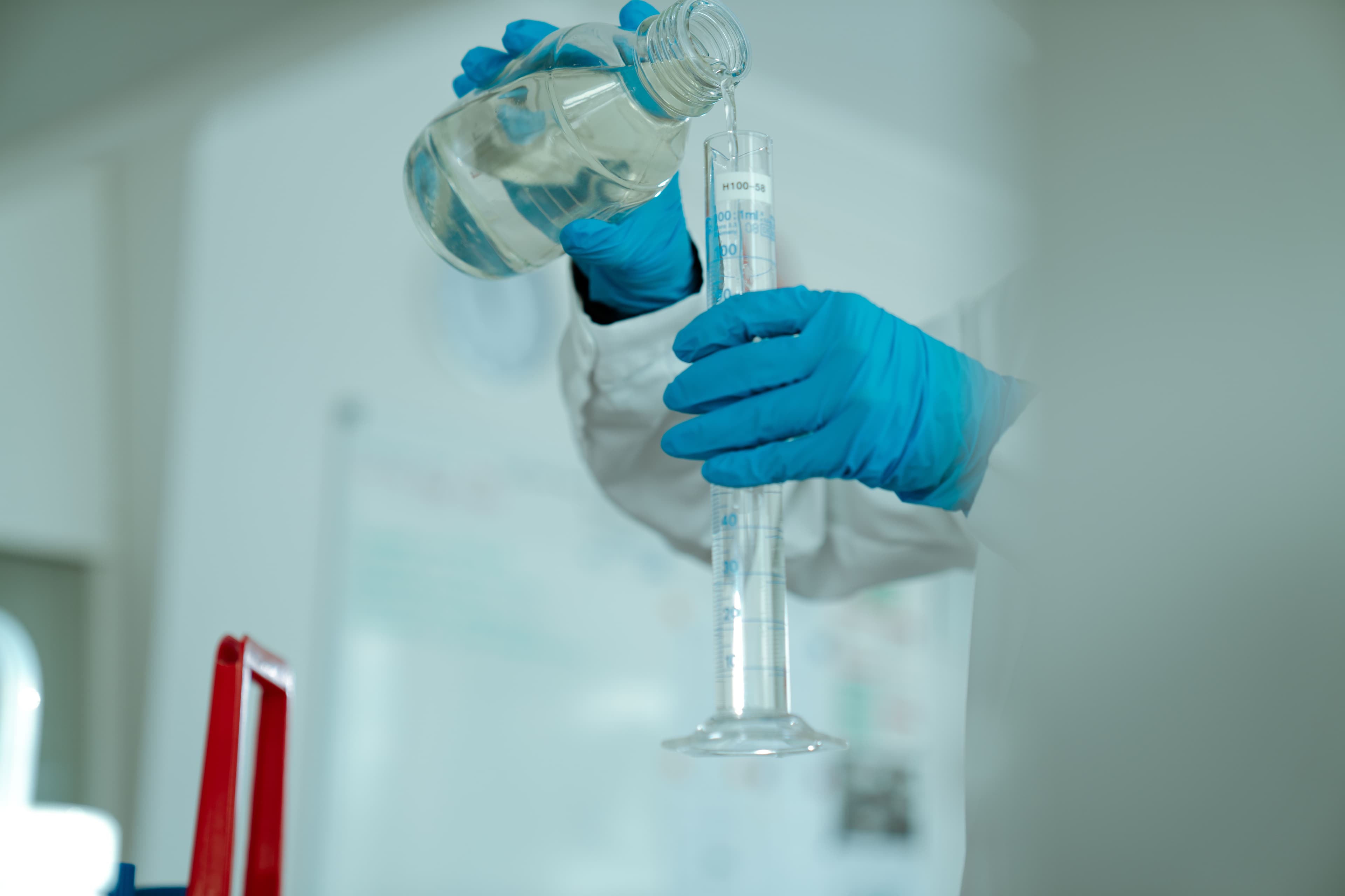 Laboritöötaja valab vedelikku mõõtesilindrisse, kandes siniseid kaitsekindaid. Protsess on osa veekvaliteedi analüüsist, mis tagab puhta ja ohutu joogivee