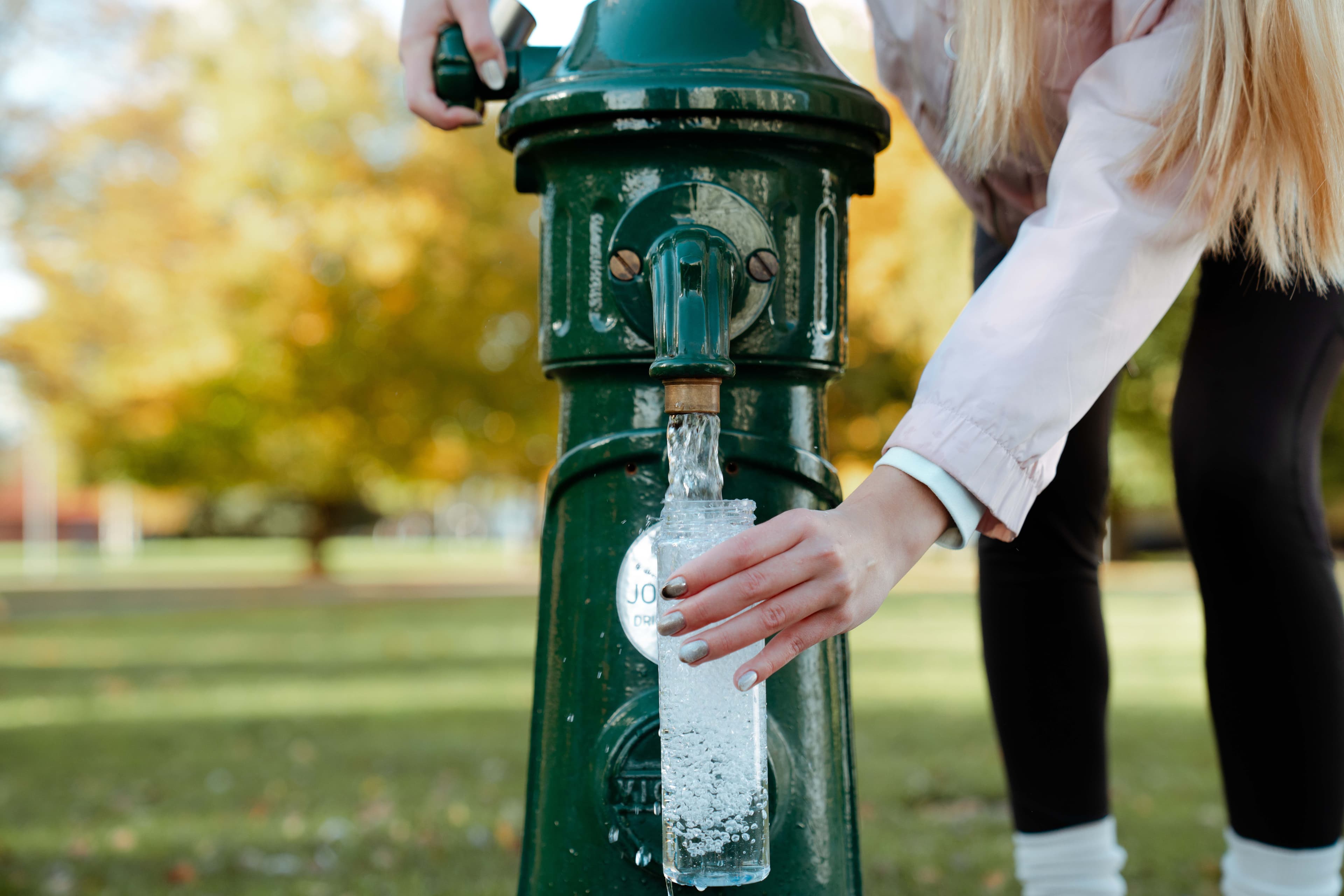 “A green metal water pump outdoors with a faucet attached. Clear water flows from the faucet into a transparent bottle held by a woman with one hand. In the background, there is green grass and trees in autumn colors