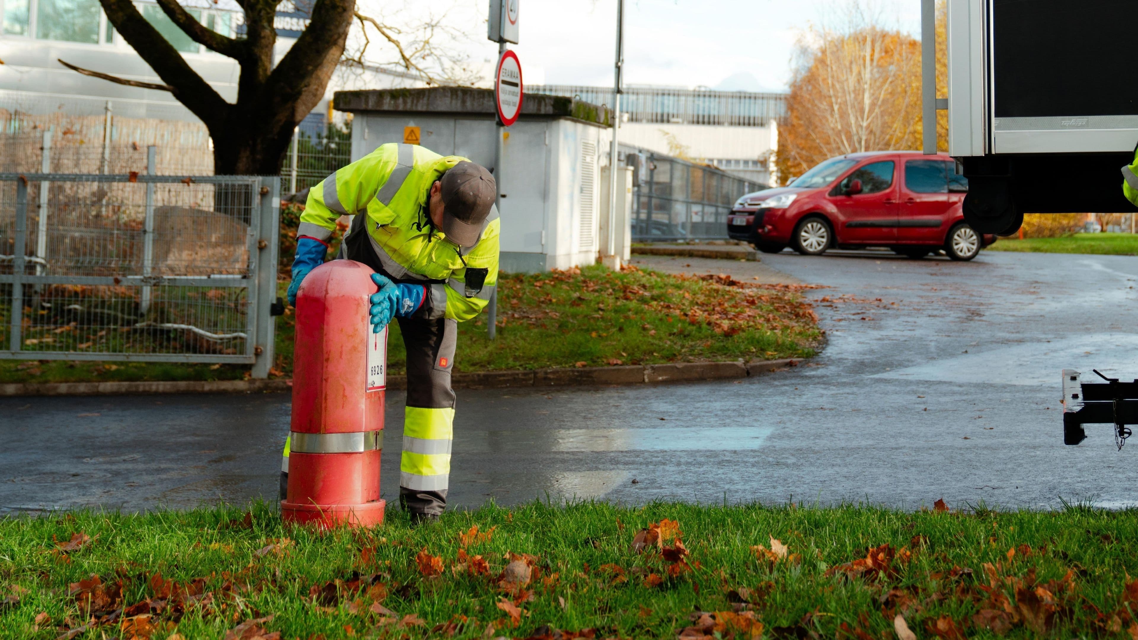 Worker in high-visibility clothing connects to a red fire hydrant to draw water, standing on a grassy area with autumn leaves.