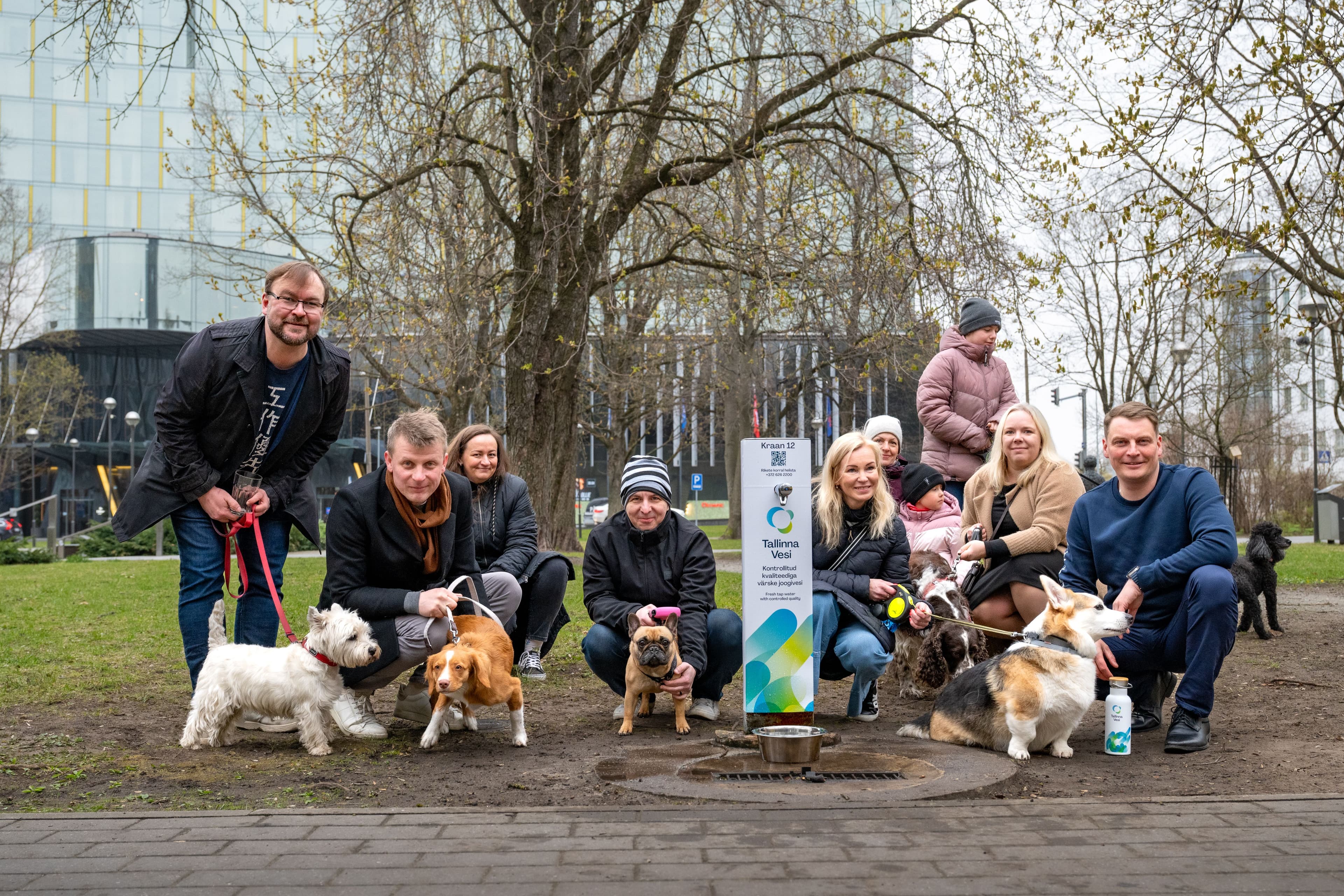 A group of people and their dogs pose next to a dog waste station in a city park.