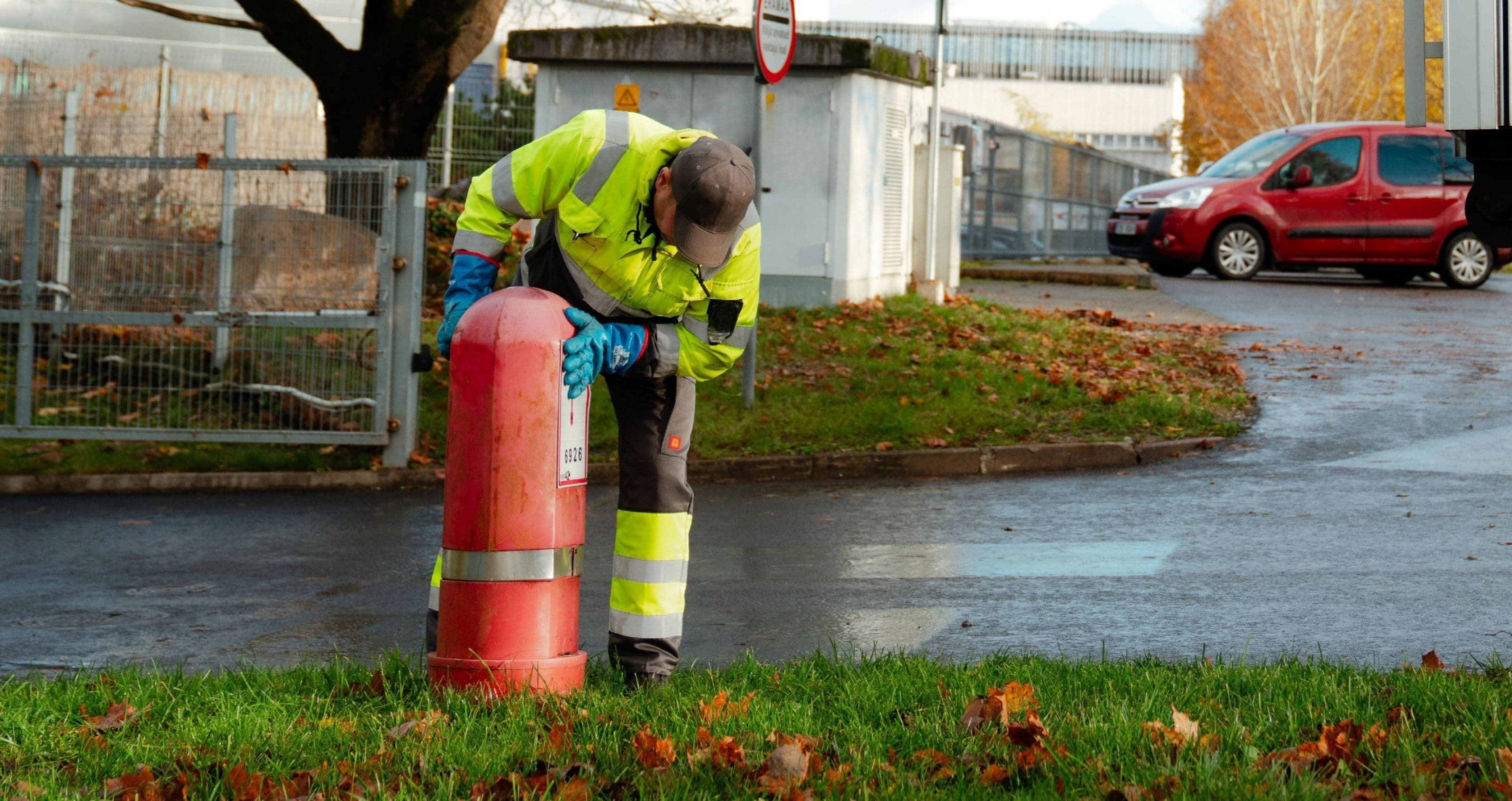 Töötaja kõrgnähtavuses tööriietes kummardub punase hüdrandi kohale, et võtta vett. Hüdrant asub rohelisel murul, kus on laiali sügislehed. Taustal on märg asfalttee, punane sõiduauto, metallist aiad ja hooned. 