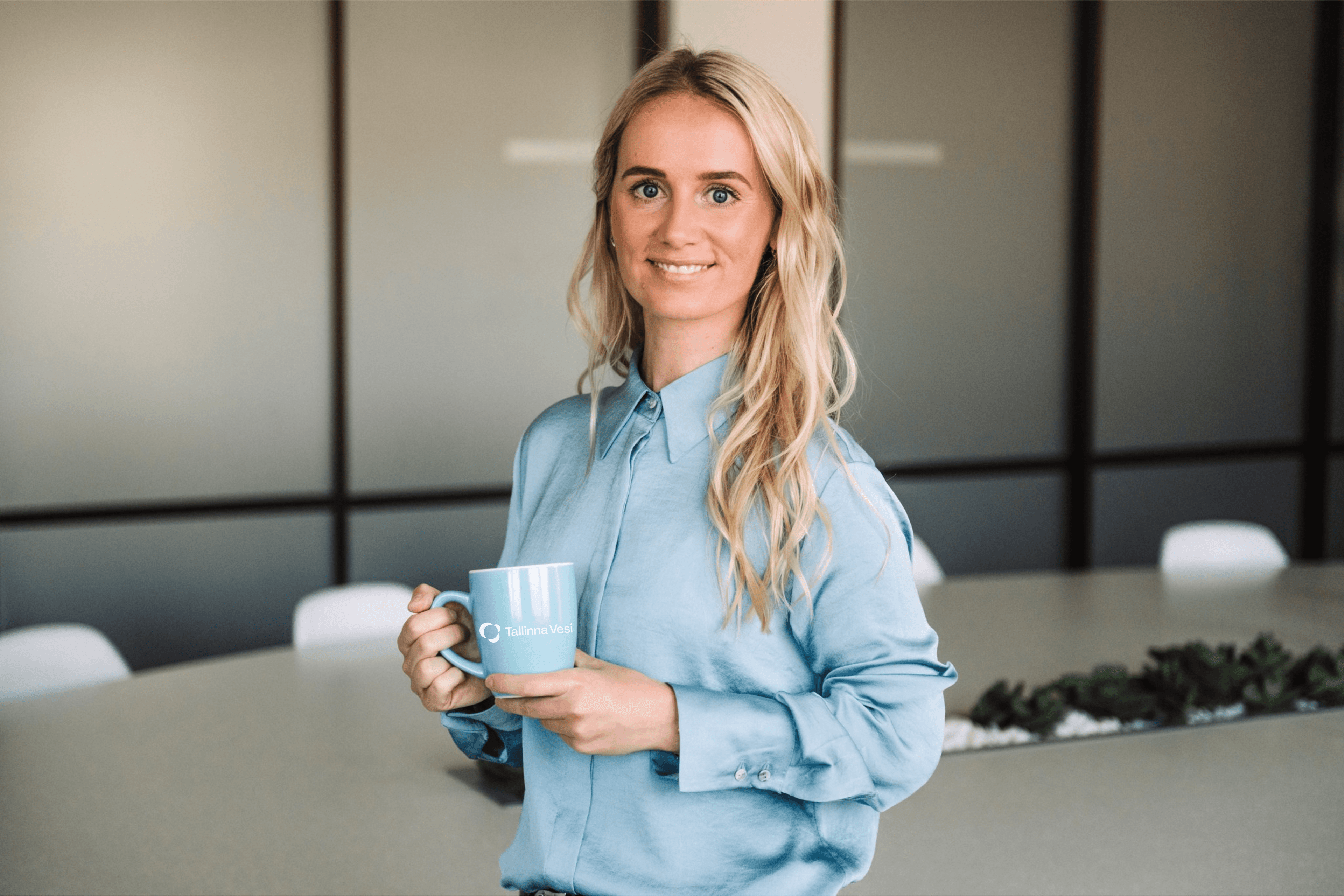Young blonde woman in blue shirt holds a light blue mug in a modern office.