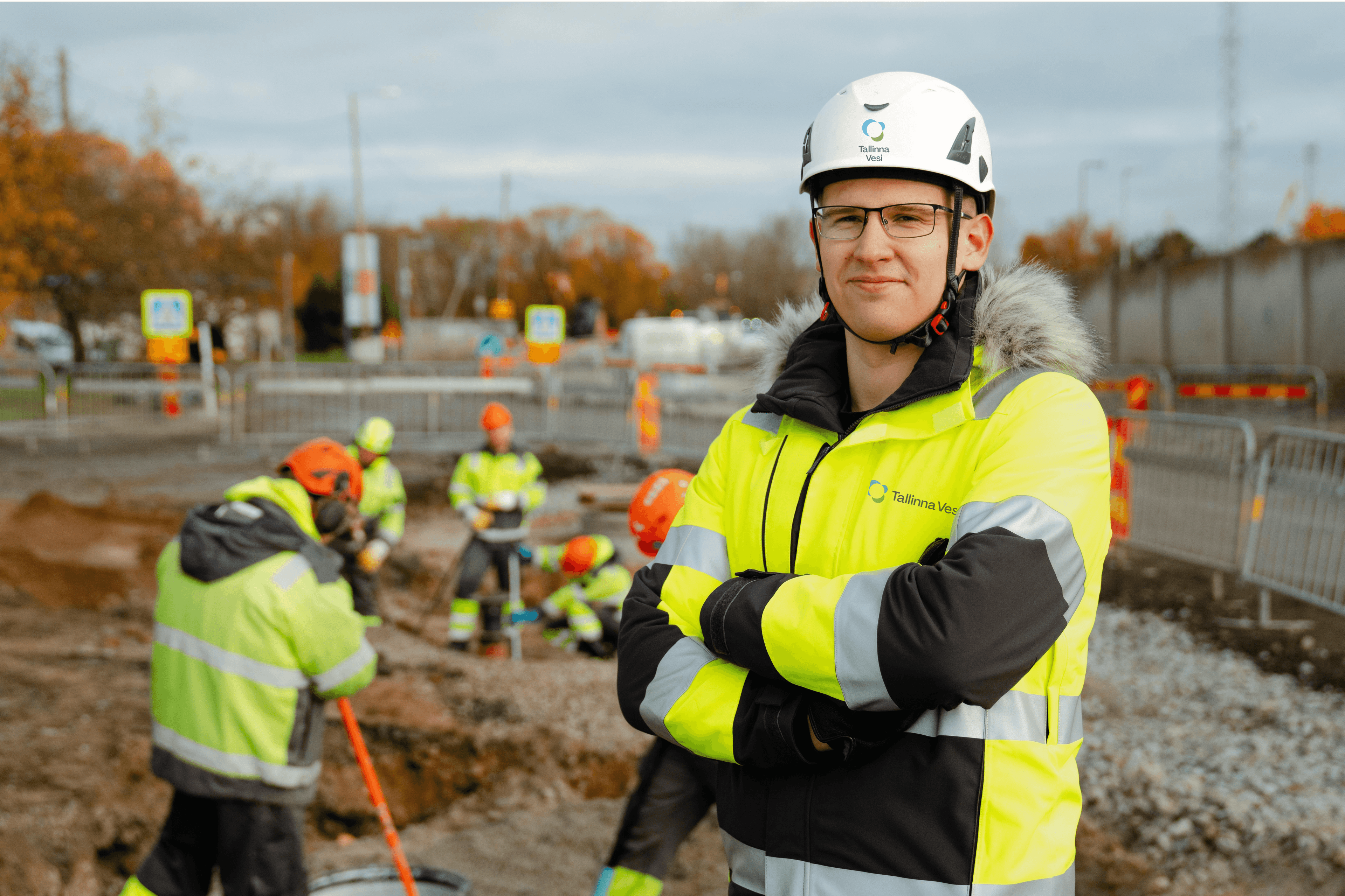 Man in white hard hat and yellow safety jacket stands with crossed arms at a construction site.