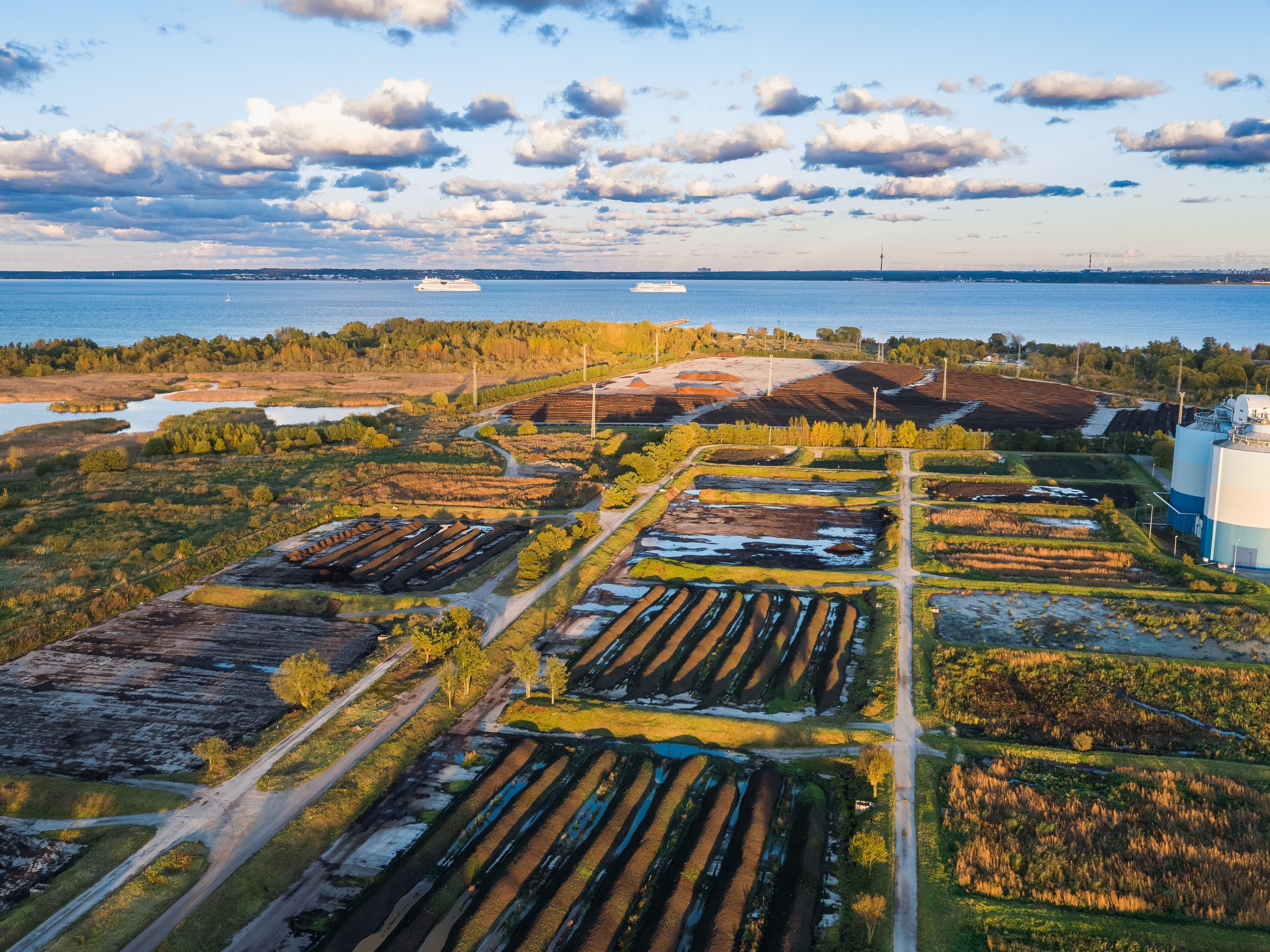 Aerial view of composting fields at Paljassaare wastewater treatment plant near the sea, with rows of dark soil, green patches, and two ships on the horizon under a partly cloudy sky.